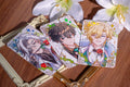 Three illustrated character cards on a wooden surface with flowers.