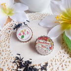 Two decorative bottle caps with floral designs on a lace doily with flowers and tea leaves.